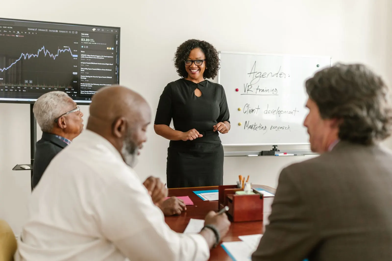 A facilitator presenting a training agenda on a whiteboard to three colleagues seated at a meeting table