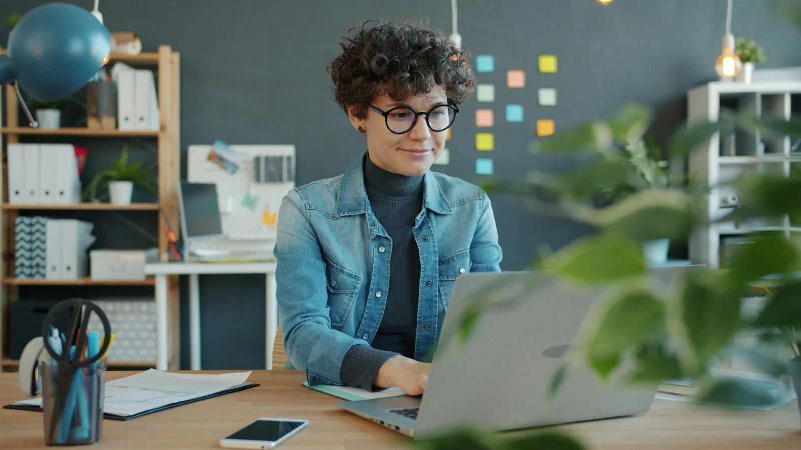 A professional with glasses concentrating on work at a laptop beside her notes