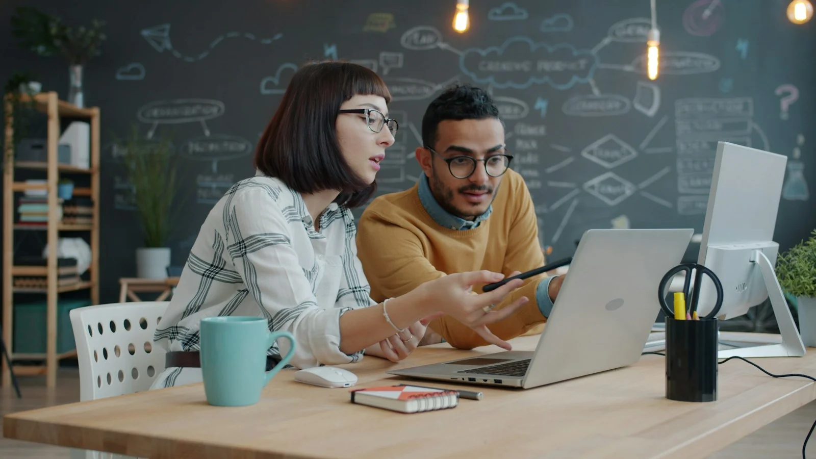 Two colleagues discussing work together with a laptop, notebook, and pen